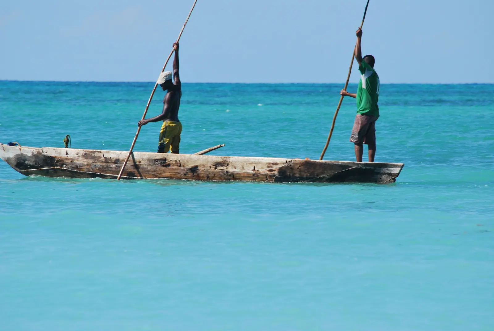 Two fishers poling a wooden canoe across shallow turquoise water